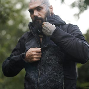 May include: Black hooded jacket featuring a dragon pattern on the front. The jacket has a zip and side pockets. The person is wearing the jacket outdoors.