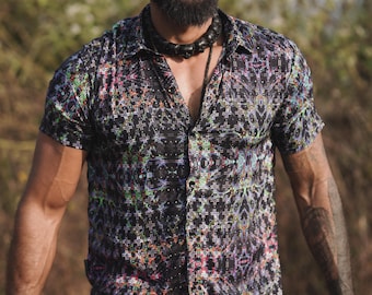 May include: A man wearing a black short-sleeved button-down shirt with a colorful geometric pattern. He has a beard, a black beaded necklace, and a large black hoop earring in his left ear.