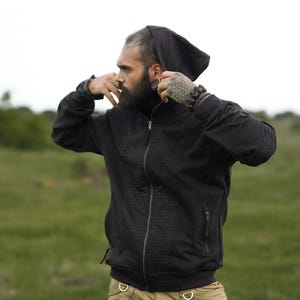 Puede incluir: Una sudadera con capucha negra con cierre de cremallera y un patrón sutil. La sudadera la lleva puesta una persona con barba y tatuajes.