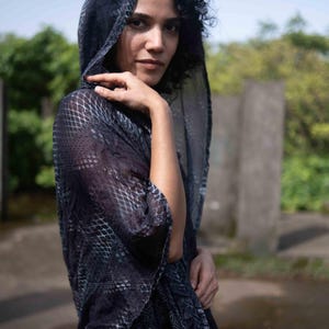 Pode incluir: Um xale com capuz azul marinho escuro com um padrão geométrico e texturizado. O xale é drapeado sobre os ombros e a cabeça, criando uma silhueta fluida. O tecido parece leve e semitransparente.