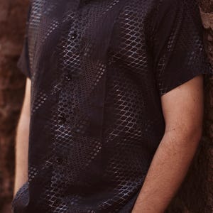 May include: A black short-sleeved button-down shirt with a geometric pattern. The shirt has a subtle sheen and a relaxed fit.