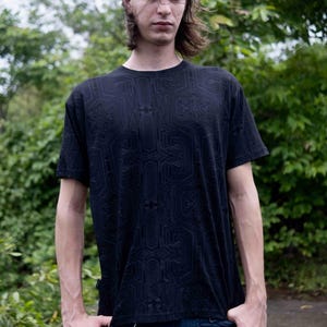 Puede incluir: Camiseta negra de manga corta con un diseño geométrico de placa de circuito. La camiseta se combina con pantalones azul oscuro. La persona está de pie al aire libre con follaje verde en el fondo.