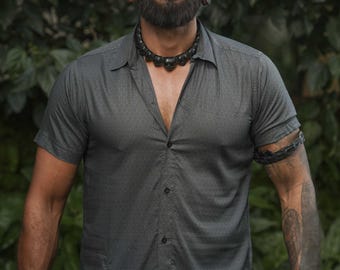 May include: A dark gray short-sleeved button-down shirt with a subtle micro-pattern, featuring a classic collar and a straight hem. The shirt is worn with a black beaded necklace, dark wrist cuffs, and a dark arm band on the left bicep.