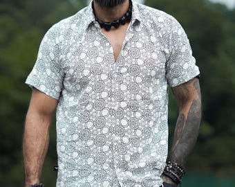 May include: A man with a shaved head and a beard is wearing a white short-sleeved shirt with a black geometric pattern. He is also wearing black shorts and several bracelets on his wrists.