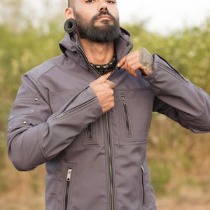 May include: A gray softshell jacket with a zipper closure and multiple pockets. The jacket has a hood and a black zipper pull.