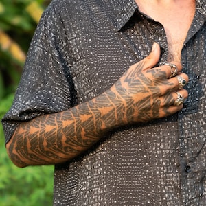 Puede incluir: Una persona lleva una camisa negra de botones con estampado de cocodrilo blanco. La camisa tiene cuello y mangas largas. El brazo de la persona está extendido, mostrando un tatuaje geométrico negro.