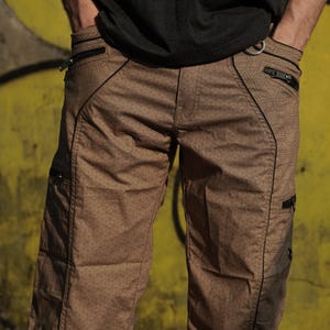 Op de afbeelding: Bruine cargo shorts met een geometrisch patroon en zwarte bies. De shorts hebben meerdere zakken met ritsen en een trekkoord aan de onderkant.