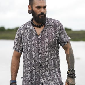 Pode incluir: Um homem vestindo uma camisa de botões de manga curta com estampa de cobra em preto e branco. Ele está vestindo calças pretas e tem barba e vários piercings nas orelhas.