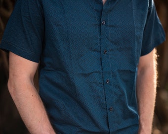 Può includere: Una camicia a maniche corte abbottonata di colore blu-verde con un motivo geometrico. La camicia è sbottonata al collo e le maniche sono arrotolate.