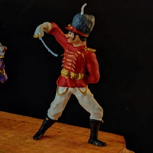 May include: A gray rat wearing a green and purple cape and a gold crown is holding a sword and facing a red and white toy soldier. The soldier is holding a white string in his right hand.
