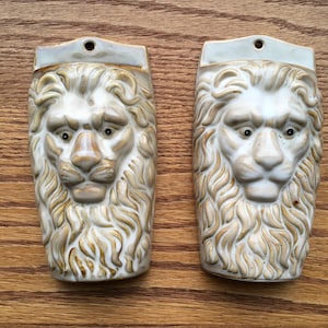 May include: Two ceramic lion head wall sculptures with a beige and brown glaze. The lion heads are facing forward with their mouths closed.