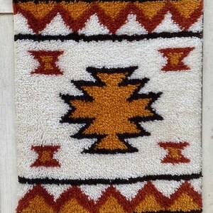 May include: A rectangular, textured rug with a geometric design. The rug features a white background with a central orange and black pattern. The border and other design elements are in shades of orange and brown.