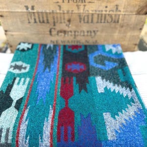 May include: A folded textile featuring a vibrant geometric pattern in shades of teal, royal blue, deep red, black, and white. The design consists of vertical bands with repeating angular shapes and serrated edges, creating a bold, multi-colored visual texture.