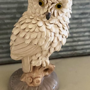 Puede incluir: Una figurita de búho de cerámica blanca con ojos amarillos, posada en una rama de árbol marrón. Las plumas del búho están esculpidas para parecer escamas.