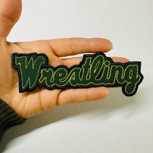 May include: A green and gold embroidered patch with the word "Wrestling" in a cursive font. The patch has a black border and is likely for a jacket or bag.