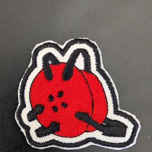 May include: Embroidered patch featuring a red design with black accents and a white border. The patch has a unique shape and a textured appearance, suitable for application on clothing or accessories. The design includes two red spheres and black lines.