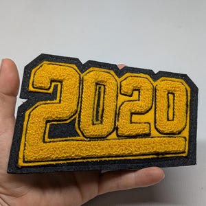 May include: A yellow and black felt patch with the year "2020" stitched in a varsity letter style. The numbers are yellow with a black outline and sit on a black background. The patch is rectangular and has a glittery border.