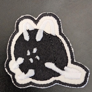 May include: Black and white embroidered patch of a cartoon character with a round head and a white outline.