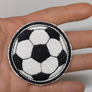 May include: Round embroidered patch depicting a football. The design features the classic black and white panels of a football, with a detailed black border. The patch is suitable for application on clothing or accessories.