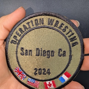 May include: A green, circular patch with black text that reads "Operation Wrestling San Diego Ca 2024". The patch is surrounded by small embroidered flags of the United Kingdom, the United States, Canada, and France.