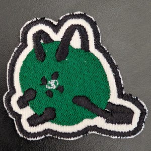 May include: Green embroidered iron-on patch of a cartoon cat with black outlines and white stitching.