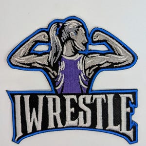 May include: An embroidered patch with a blue outline. The patch features a female figure with her arms raised in a flexing pose. The figure is wearing a purple wrestling singlet. The text "WRESTLE" is below the figure in white letters with a black outline.