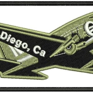 May include: Embroidered patch featuring a stylized airplane in black and olive green, with the text "San Diego, Ca" in white. The patch is rectangular with a black border, suitable for application on clothing or accessories.