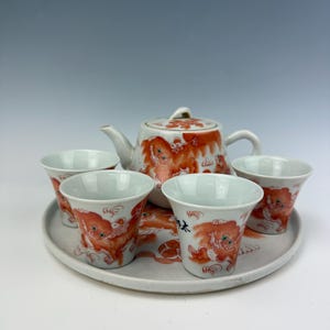 May include: A white ceramic tea set consisting of a teapot and four small cups arranged on a matching round tray. Each piece features hand-painted orange lion-like creatures with green eyes and black ink calligraphy characters on the exterior surfaces.