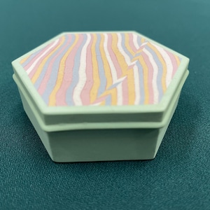 May include: A hexagonal trinket box with a light green base and lid. The lid features a wavy pattern in pastel colors of pink, yellow, blue, and white, with a lightning bolt design. The box is on a green surface.