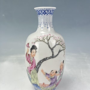 Può includere: Un vaso di porcellana bianca con bordo e base blu a motivi. Il vaso presenta una scena dipinta a mano di figure sotto un albero in fiore, con fiori rosa e una farfalla. Le figure indossano abiti colorati.