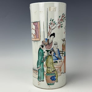 May include: A cylindrical ceramic vase with a white base featuring a hand-painted scene of figures in traditional attire gathered around a table. The artwork includes green, blue, brown, and red accents, depicting people, a tree, and a window.