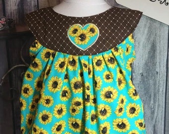 Peut inclure: Un barboteuse turquoise pour bébé avec un imprimé tournesol. La barboteuse présente un col marron avec un motif de losanges blancs et une application de tournesol en forme de cœur. Le motif comprend des tournesols jaunes avec des centres noirs.