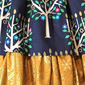 Peut inclure: Gros plan sur un tissu bleu marine avec un motif d'arbre blanc orn&eacute; de fleurs bleues, roses et vertes et de d&eacute;tails dor&eacute;s en forme d'&eacute;toiles. Une volants dor&eacute;s textur&eacute;s avec des motifs de flocons de neige et de cachemire ajoute une couche contrastante.