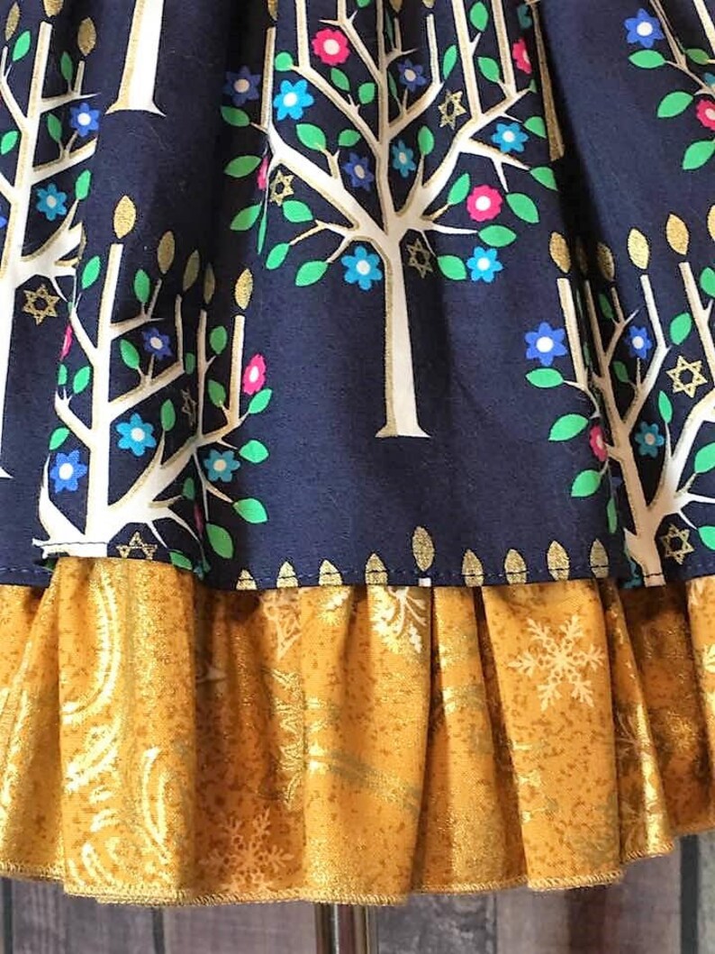 Peut inclure: Gros plan sur un tissu bleu marine avec un motif d'arbre blanc orn&eacute; de fleurs bleues, roses et vertes et de d&eacute;tails dor&eacute;s en forme d'&eacute;toiles. Une volants dor&eacute;s textur&eacute;s avec des motifs de flocons de neige et de cachemire ajoute une couche contrastante.