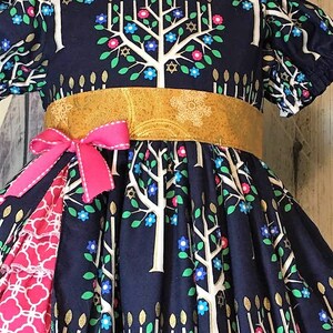 Peut inclure: Robe bleu marine orn&eacute;e d'un motif d'arbre de vie avec des branches blanches, des accents dor&eacute;s en forme de bougies et des fleurs color&eacute;es. La robe a une ceinture dor&eacute;e, un n&oelig;ud rose et un volant &agrave; motifs rouges et blancs.