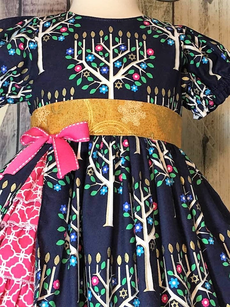 Peut inclure: Robe bleu marine orn&eacute;e d'un motif d'arbre de vie avec des branches blanches, des accents dor&eacute;s en forme de bougies et des fleurs color&eacute;es. La robe a une ceinture dor&eacute;e, un n&oelig;ud rose et un volant &agrave; motifs rouges et blancs.