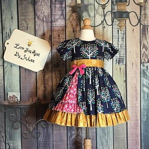 Peut inclure: Robe bleu marine &agrave; motif d'arbres blancs, ceinture dor&eacute;e et n&oelig;ud rose. La jupe pr&eacute;sente un panneau &agrave; motifs roses et blancs et un ourlet &agrave; volants dor&eacute;s. L'&eacute;tiquette indique "Love You Most By So Ann."