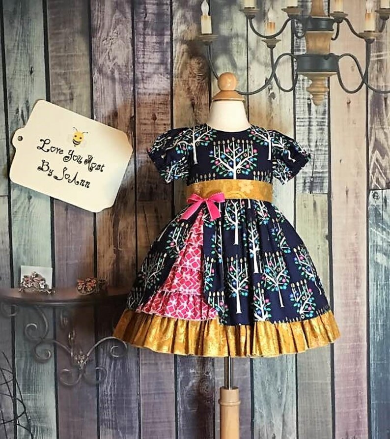 Peut inclure: Robe bleu marine &agrave; motif d'arbres blancs, ceinture dor&eacute;e et n&oelig;ud rose. La jupe pr&eacute;sente un panneau &agrave; motifs roses et blancs et un ourlet &agrave; volants dor&eacute;s. L'&eacute;tiquette indique "Love You Most By So Ann."