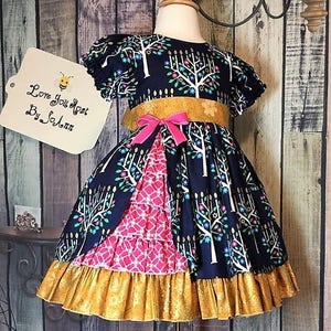 Peut inclure: Robe bleu marine avec un motif d'arbres, une ceinture dor&eacute;e et un n&oelig;ud rose. La robe a un panneau &agrave; motifs roses et blancs et un ourlet &agrave; volants dor&eacute;s. L'&eacute;tiquette indique "Love You Most By Solann".