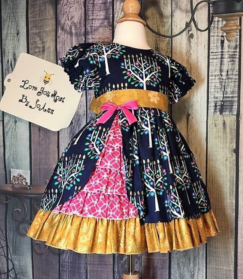 Peut inclure: Robe bleu marine avec un motif d'arbres, une ceinture dor&eacute;e et un n&oelig;ud rose. La robe a un panneau &agrave; motifs roses et blancs et un ourlet &agrave; volants dor&eacute;s. L'&eacute;tiquette indique "Love You Most By Solann".