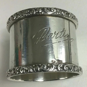 Puede incluir: Un servilletero plateado con el nombre "Berti" grabado. Presenta patrones ornamentales en espiral en los bordes superior e inferior. La superficie metálica pulida refleja la luz, resaltando los detalles.