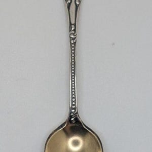 May include: A silver-toned metal spoon featuring an ornate, embossed floral pattern on the handle and a beaded border detail along the stem. The bowl of the spoon has a smooth, reflective surface, and the handle includes an engraved script letter.