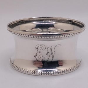 May include: A silver napkin ring with a wide, cylindrical form. The top and bottom edges have a beaded design, and the centre features an engraved "EW" monogram. The polished surface reflects light, creating a bright appearance.