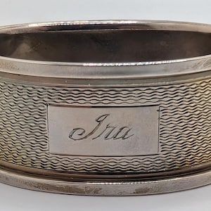 May include: A silver napkin ring with a textured, wave-like pattern around the body. The name "Ira" is engraved on a rectangular plaque. The ring has a simple, elegant design.