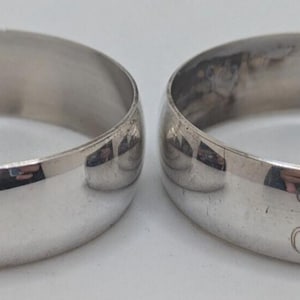 May include: Two polished silver-toned metal bands, each featuring a smooth, rounded exterior surface. A single, elegant, cursive letter J is engraved on the front centre of each ring, providing a subtle, personalised detail on the reflective surface.