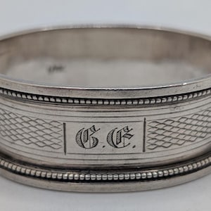 Puede incluir: Un servilletero plateado con un diseño detallado. El anillo presenta un patrón texturizado y está grabado con las iniciales "G.E." en una fuente decorativa. Los bordes están adornados con un borde de cuentas, lo que aumenta su atractivo clásico.