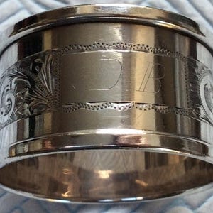 Könnte beinhalten: Ein silberner Serviettenring mit gravierten Blumen- und Scrollmustern. Der Ring hat eine rechteckige Kartusche mit den Initialen "DB" in einer Serifenschrift. Die Kanten sind glatt und poliert und reflektieren das Licht.