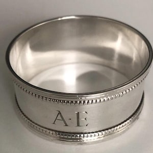 May include: A silver napkin ring with a polished finish. The ring has a decorative beaded border and engraved initials "A.E." in the center. The ring is circular and appears to be made of metal.