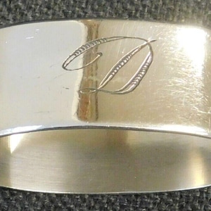Puede incluir: Un servilletero plateado con un acabado pulido. El anillo presenta una letra "D" grabada en escritura cursiva. El servilletero es un diseño simple y elegante, adecuado para cenas formales.