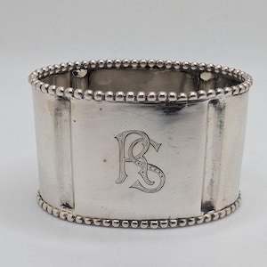 May include: A silver napkin ring with a beaded edge. The ring has a rectangular shape with a monogram of the letters "RS" engraved on the front. The ring is made of a shiny metal.